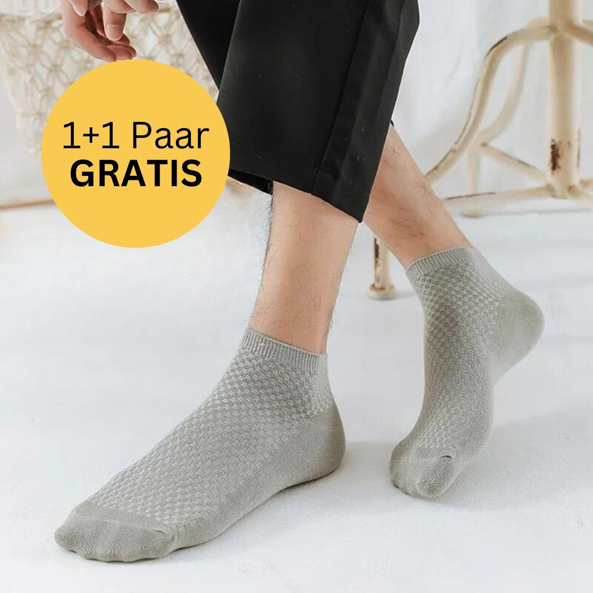 Eine Person, die hellgraue Tieberg™ Bambus Socken (Kurz) trägt, sitzt in einem Haus und legt eine Hand auf ihr Bein. In einem gelben Kreis steht "1+1 Paar GRATIS". Die Szene zeigt einen weißen Bodenbelag und helle Möbel, die die Marke Tieberg hervorheben.