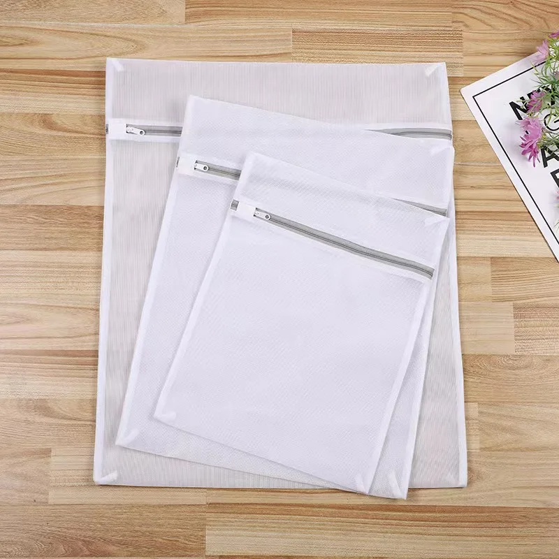 Drei weiße Tieberg Wäschenetze - für längere Haltbarkeit - mit Reißverschluss in S, M und L liegen auf Holz. Ideal für empfindliche Kleidung. Rechts oben sind teilweise ein Magazin und Blumen zu sehen.
