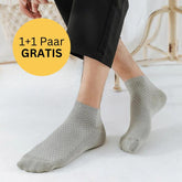 Eine Person, die hellgraue Tieberg™ Bambus Socken (Kurz) trägt, sitzt in einem Haus und legt eine Hand auf ihr Bein. In einem gelben Kreis steht "1+1 Paar GRATIS". Die Szene zeigt einen weißen Bodenbelag und helle Möbel, die die Marke Tieberg hervorheben.