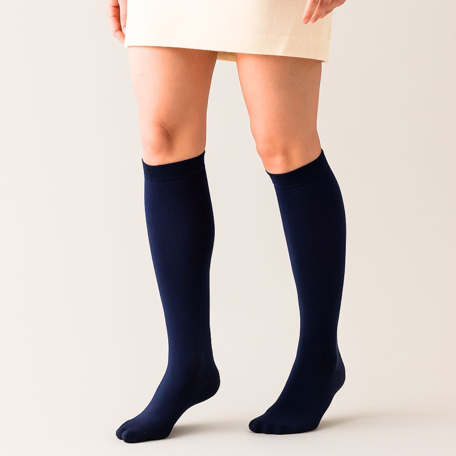 Eine Person steht vor einem hellen neutralen Hintergrund und trägt kniehohe marineblaue Tieberg™️ Vitalsocks - Spürbar leichter leben und einen cremefarbenen Rock.