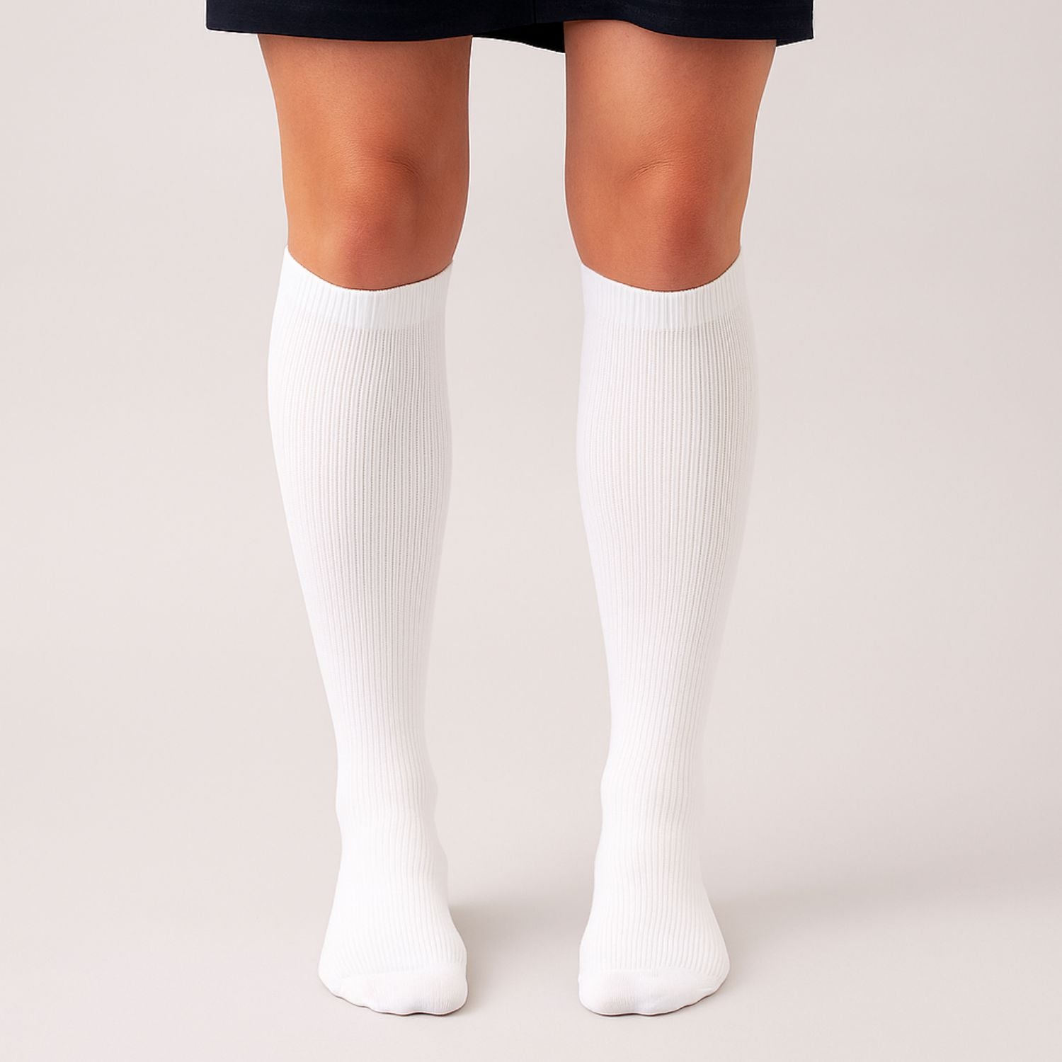 Jemand steht mit zusammengelegten Füßen auf einer hellen Fläche, trägt einen dunklen Rock und kniehohe gerippte weiße Vitalsocks von Tieberg™️ und blickt nach vorne.